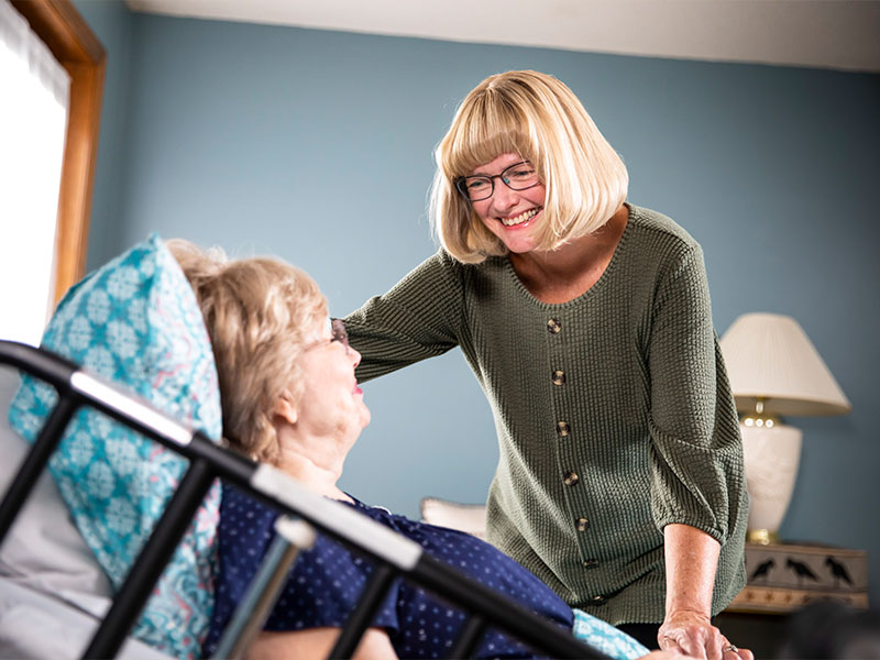 Daughter stands and smiles while speaking with aging mother in bed