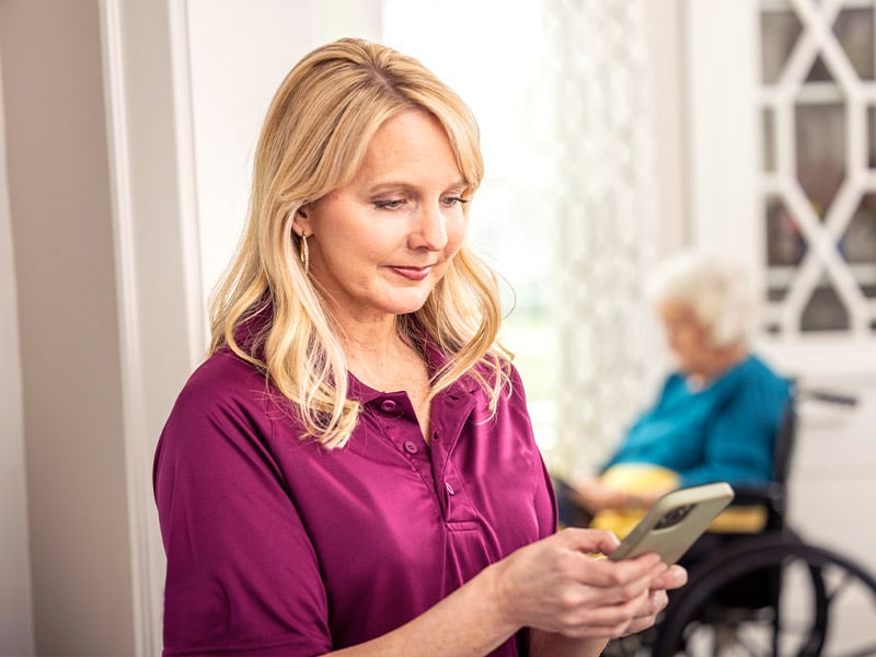 Care Professional checks care notes on her mobile phone with aging woman in background